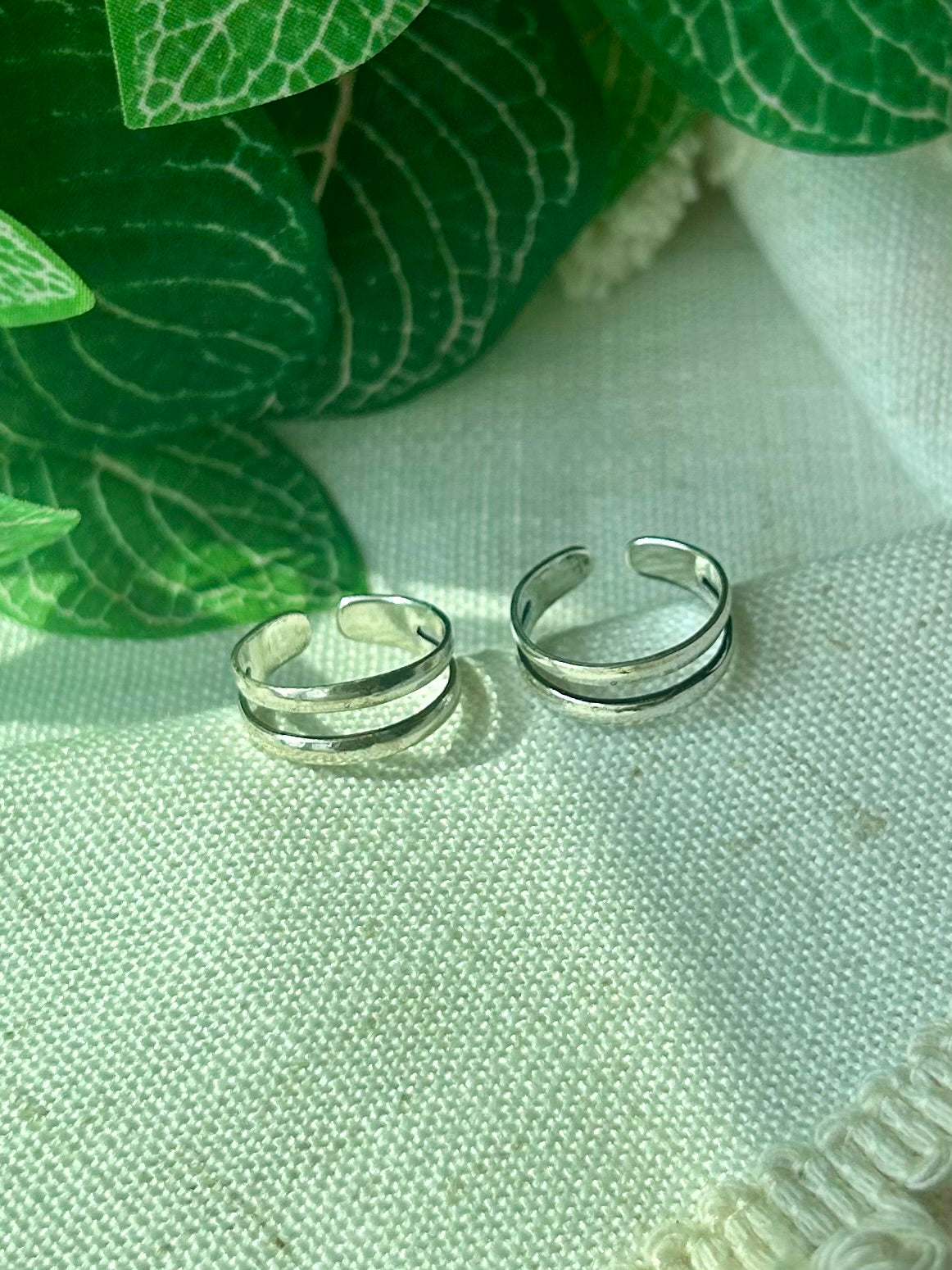Two slim silver band open-toe rings resting on a textured off-white fabric surface, with green leaves in the background.