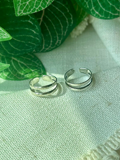Two slim silver band open-toe rings resting on a textured off-white fabric surface, with green leaves in the background.