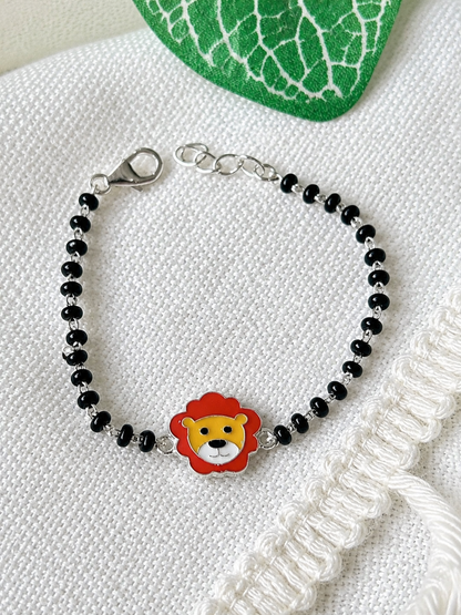 A beaded bracelet with small black beads, a silver lobster clasp, and a central lion face charm with red, yellow, and white enamel, displayed on a textured white fabric background.