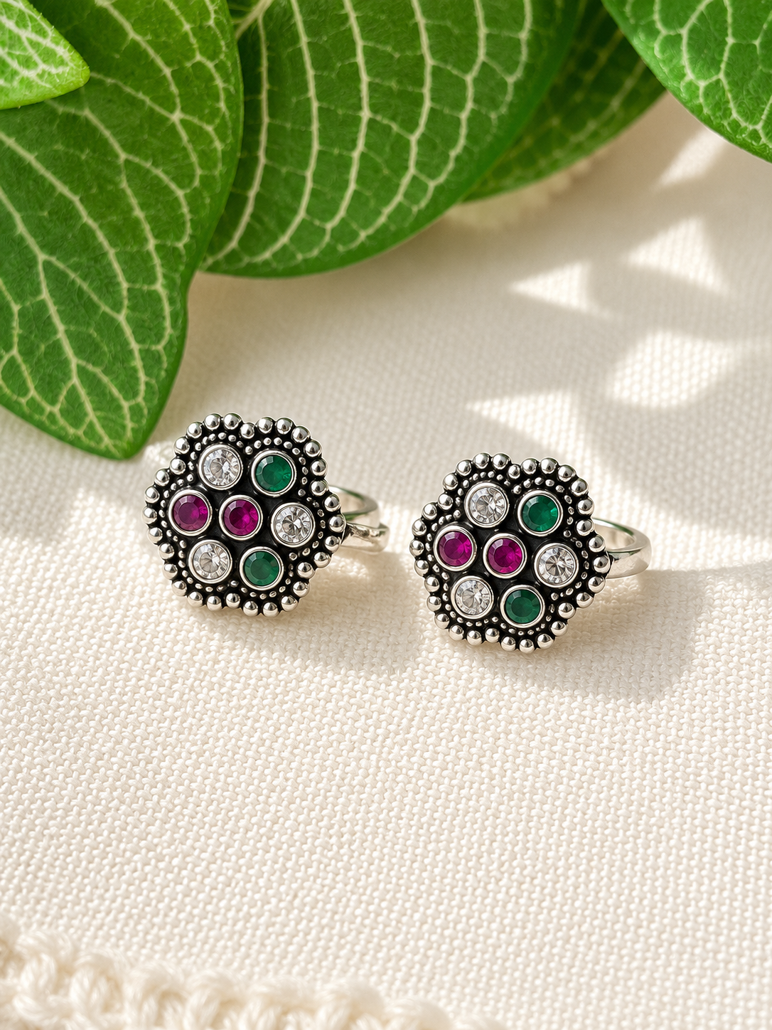 A pair of silver-toned cluster earrings with green and pink gemstones and clear stones, displayed on a textured cream fabric with green leaves in the background.