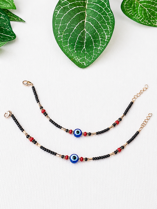 Two beaded bracelets with black beads, red accent beads, and evil eye charms, displayed on a white textured background with green leaves.