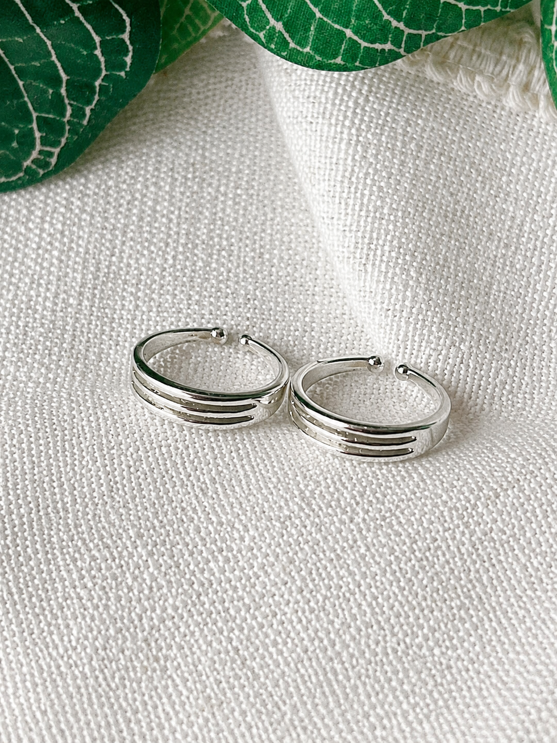 A pair of silver hoop earrings with three parallel grooved bands, resting on a textured white fabric background with green leaves in the corner.