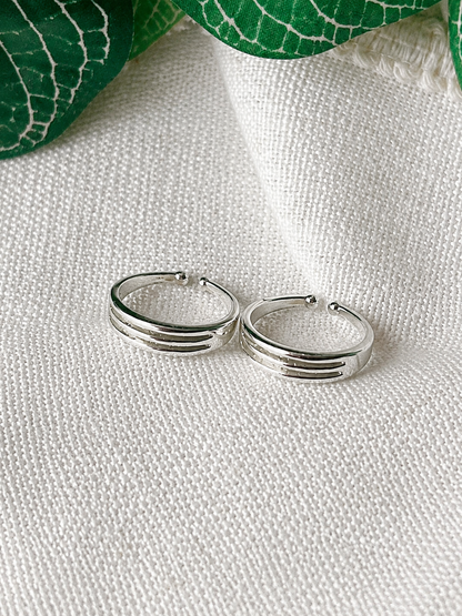 A pair of silver hoop earrings with three parallel grooved bands, resting on a textured white fabric background with green leaves in the corner.