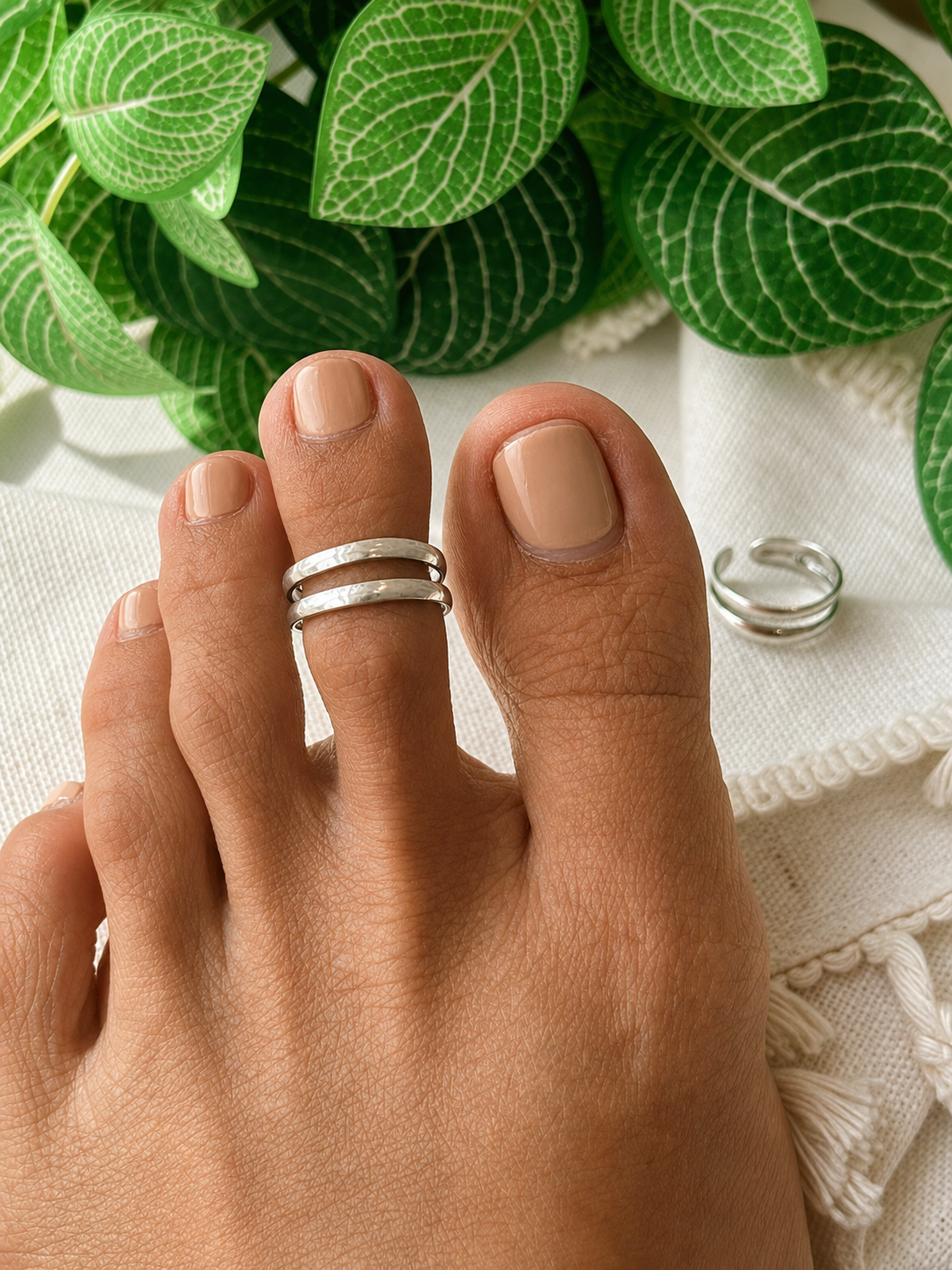 A close-up of a person's foot wearing two thin silver toe rings, with a matching silver ring placed beside the foot on a white textured surface and green leaves in the background.