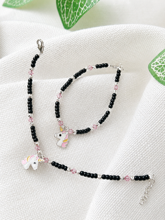A beaded bracelet and matching anklet set featuring black round beads, small pink faceted beads, silver-toned metal connectors, and white unicorn charms with pastel rainbow accents, displayed on a textured white fabric background with green leaves.