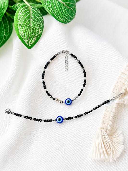 Two beaded bracelets with black beads and evil eye charms, displayed on a white textured fabric with green leaves and a cream tassel prop.