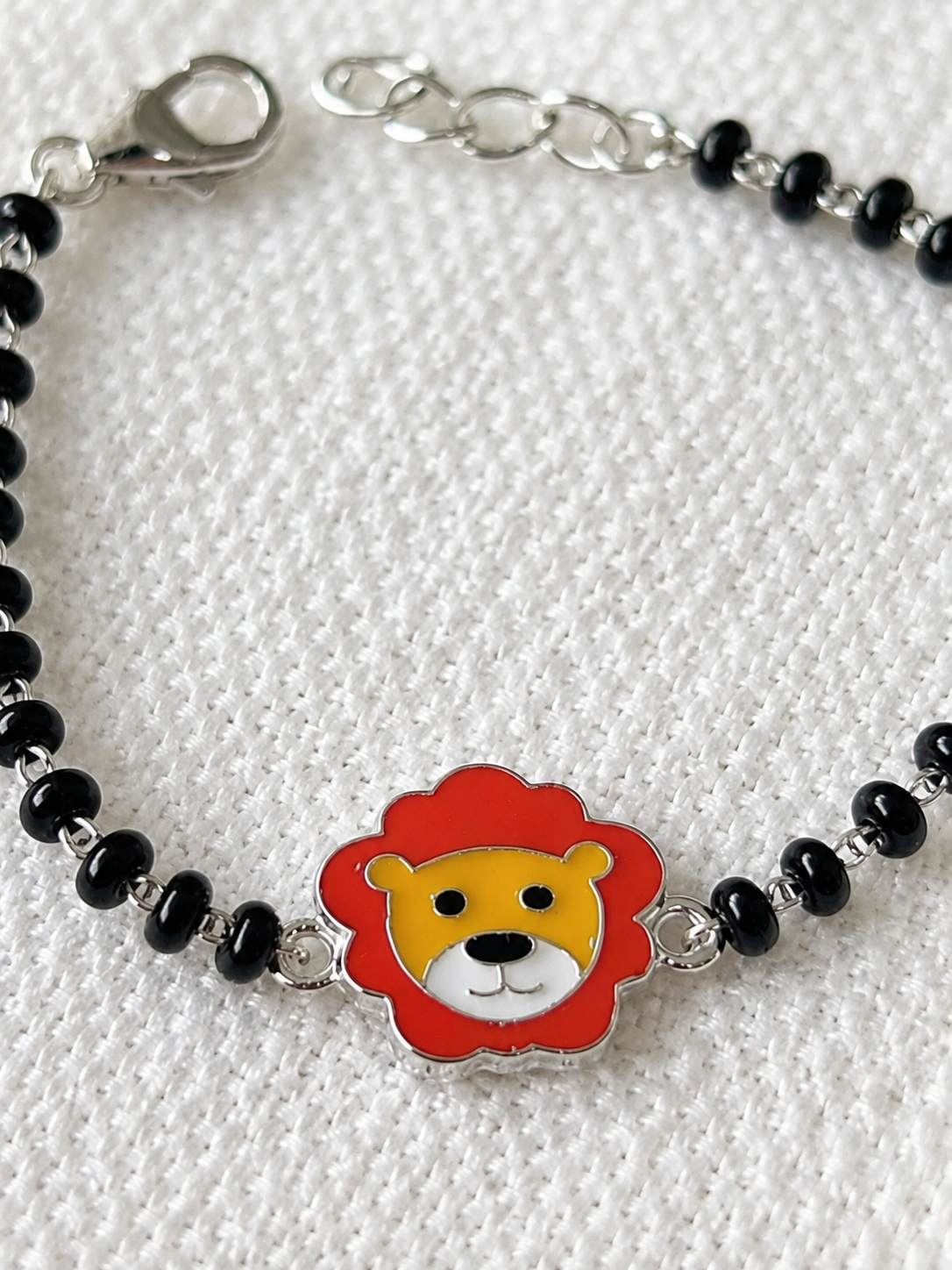 A silver beaded bracelet with a central lion head charm featuring a red mane, yellow face, and white details, displayed on a textured white fabric background.