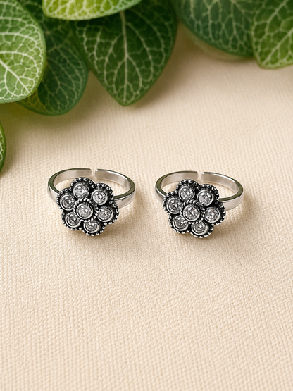 A pair of silver-toned open-shank rings with oval cluster floral motifs featuring clear stones and dark oxidized detailing, displayed on a beige textured surface with green leaves in the background.