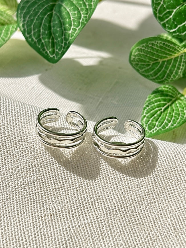 A pair of silver open-bend ring earrings with a wavy, layered design, displayed on a textured beige fabric with green leaves in the background.