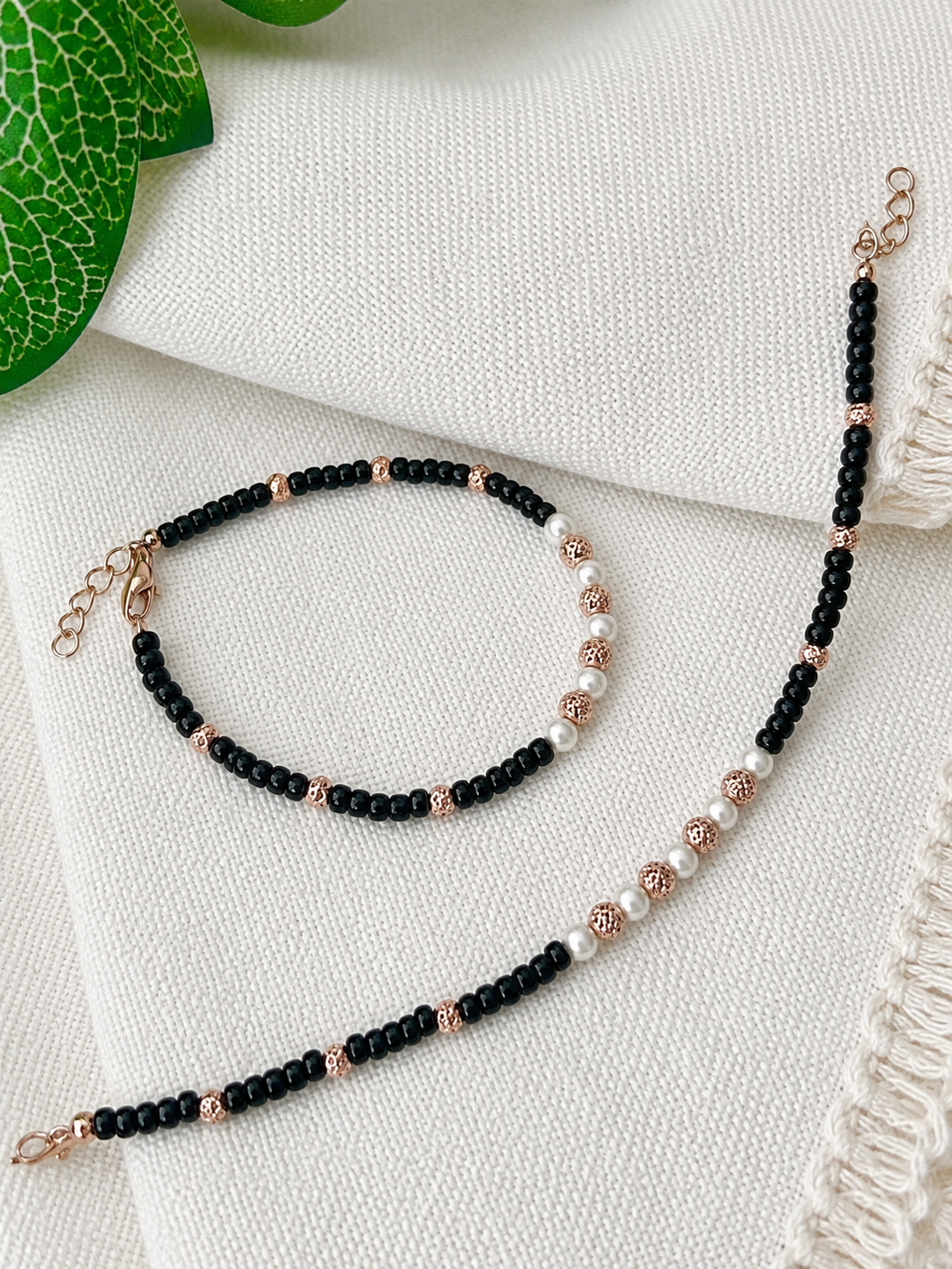 A beaded bracelet and matching anklet laid out on a textured white fabric background, featuring black round beads, white pearl-like beads, and rose gold spacer accents.