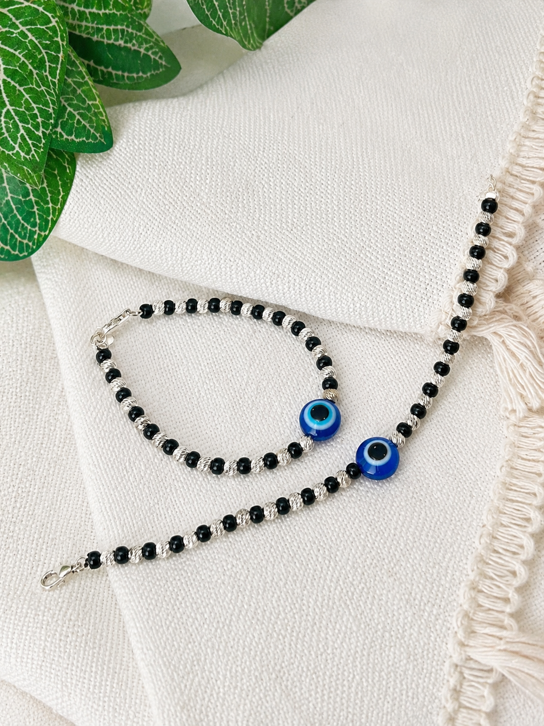 A beaded bracelet and matching anklet set with black beads, silver spacer beads, and blue evil eye charms, displayed on a textured cream fabric with green leaves in the corner.