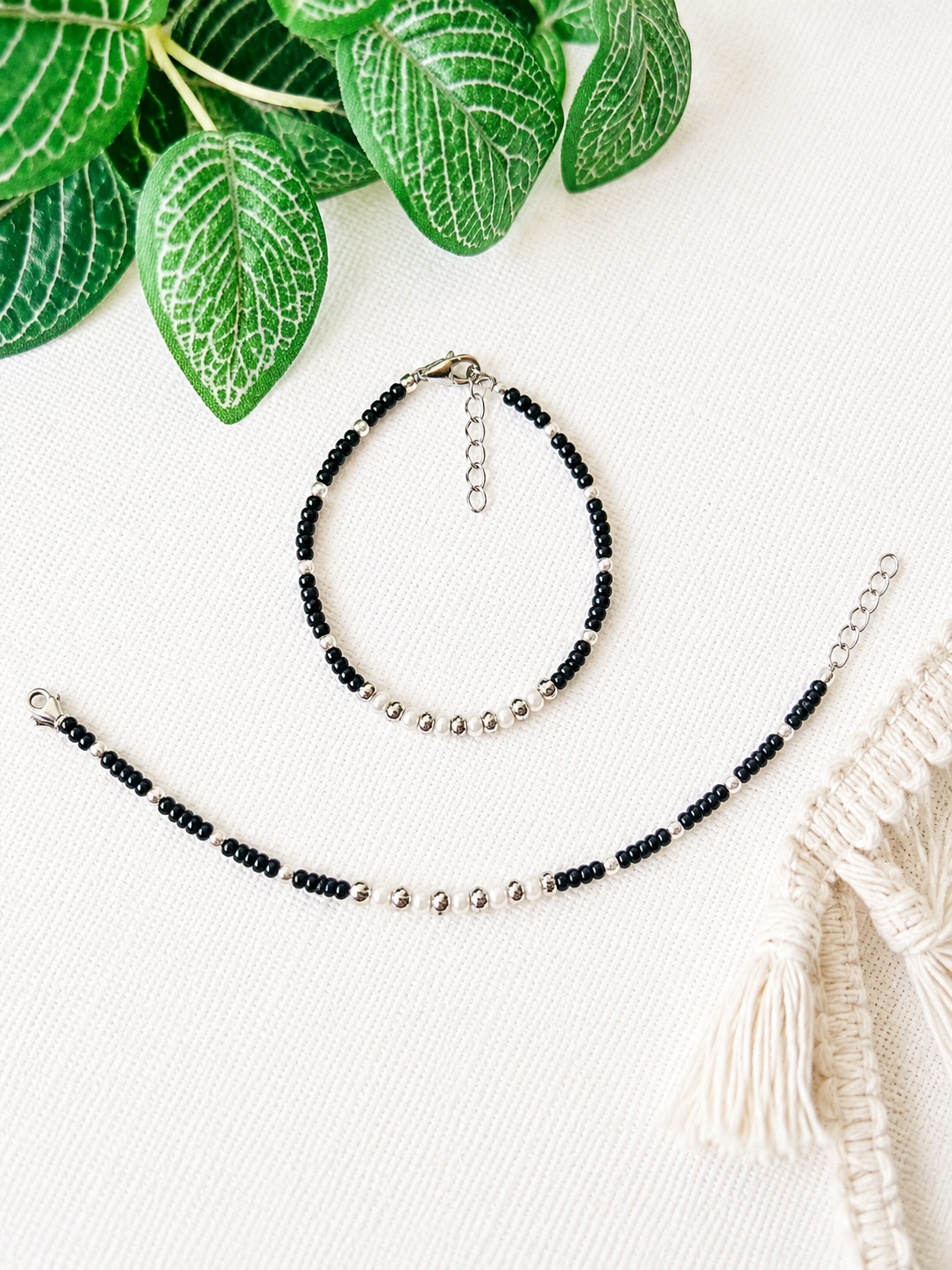 A beaded bracelet and matching anklet with black round beads and small silver metallic beads, arranged on a white textured fabric with green leaves and a cream macrame textile as props.