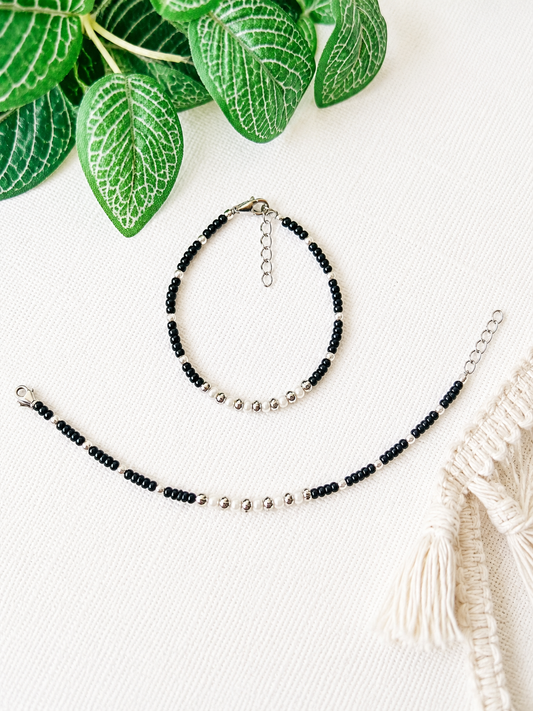 A beaded bracelet and matching anklet with black round beads and small silver metallic beads, arranged on a white textured fabric with green leaves and a cream macrame textile as props.