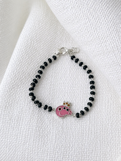A beaded bracelet with small black beads, a silver clasp, and a pink cow charm with a small gold crown, displayed on a textured white fabric background with green leaves at the top.