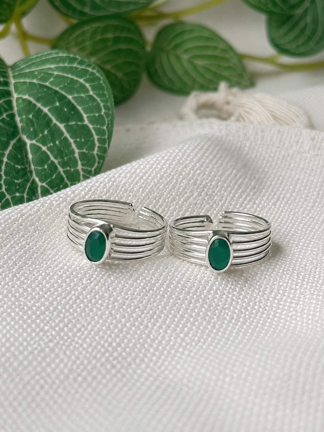 A pair of silver-toned multi-band rings with oval green stone accents, displayed on a textured white fabric with green leaves in the background.
