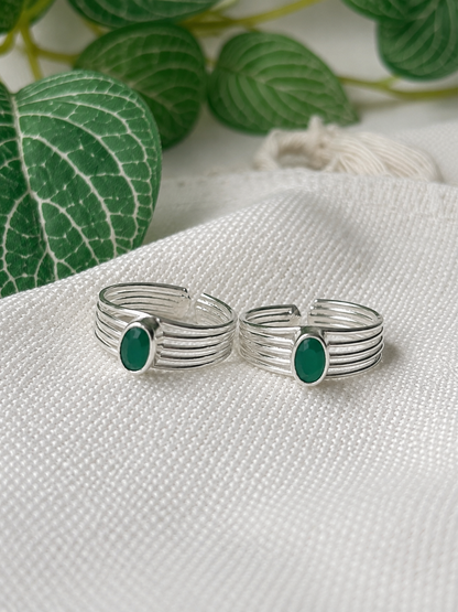 A pair of silver-toned multi-band rings with oval green stone accents, displayed on a textured white fabric with green leaves in the background.