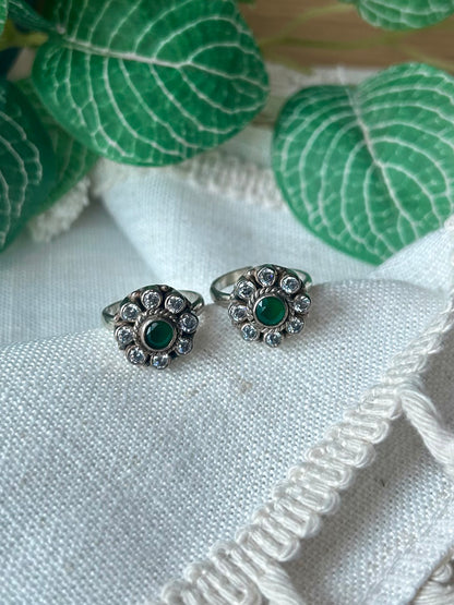 A pair of silver-toned rings with oval green gemstones surrounded by small clear stones, displayed on a textured white fabric with green leaves in the background.